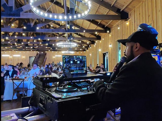 On the microphone at the booth in a barn venue with string lights and a chandelier overhead